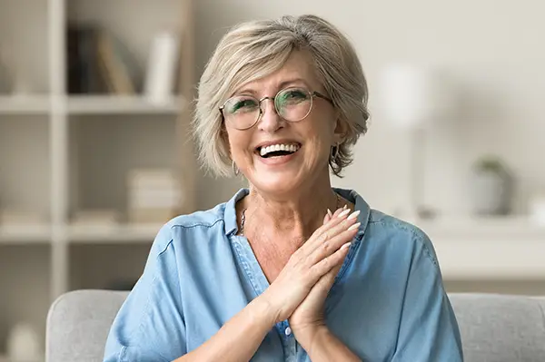 Cheerful pretty older woman in elegant glasses sitting on cozy home couch with dental implants at Fairbanks Periodontal Associates in Fairbanks, AK Cheerful pretty older woman in elegant glasses sitting on cozy home couch with dental implants at Fairbanks Periodontal Associates in Fairbanks, AK