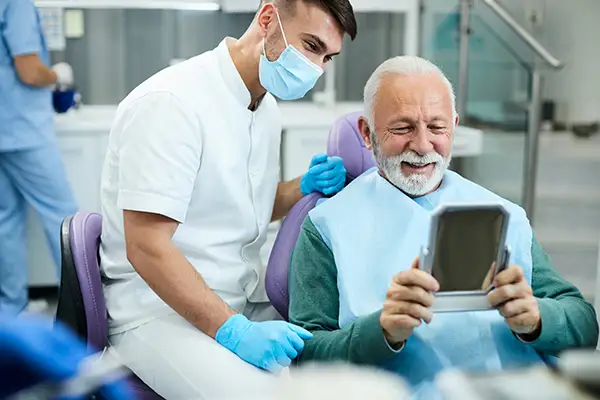 Happy, senior patient looking at himself in a mirror with dental implants at Fairbanks Periodontal Associates in Fairbanks, AK Happy, senior patient looking at himself in a mirror with dental implants at Fairbanks Periodontal Associates in Fairbanks, AK